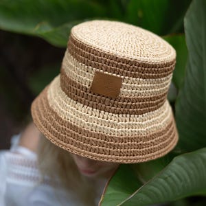 May include: A woven bucket hat with alternating bands of light beige and brown. A small, brown leather tag with text is sewn on the front. The hat is set against a backdrop of green leaves.