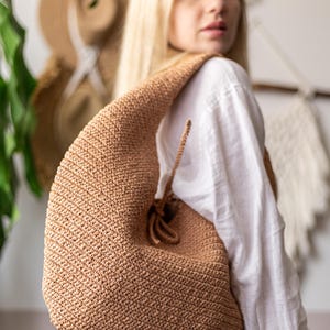 May include: A tan, crocheted hobo bag with a drawstring closure. The bag is made of a textured yarn and has a curved shape. The bag is worn over the shoulder.