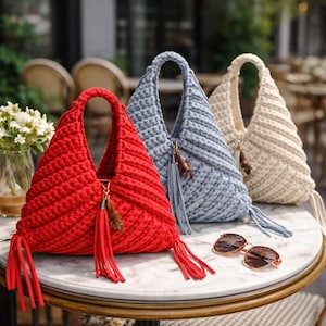 Peut inclure: Trois sacs en crochet faits à la main, rouges, bleus et beiges, sont posés sur une table en marbre. Chaque sac a un pompon unique et une anse. Des lunettes de soleil reposent sur la table. Les sacs sont exposés à l'extérieur, suggérant un thème de mode ou d'accessoires.