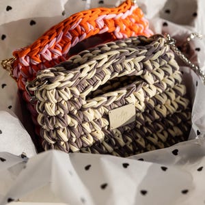 May include: Two handmade crochet bags in a gift box. One bag is a mix of beige and grey, with a silver chain strap and a small metal tag. The other bag has an orange, pink, and white handle. The gift box has a white background with black heart shapes.