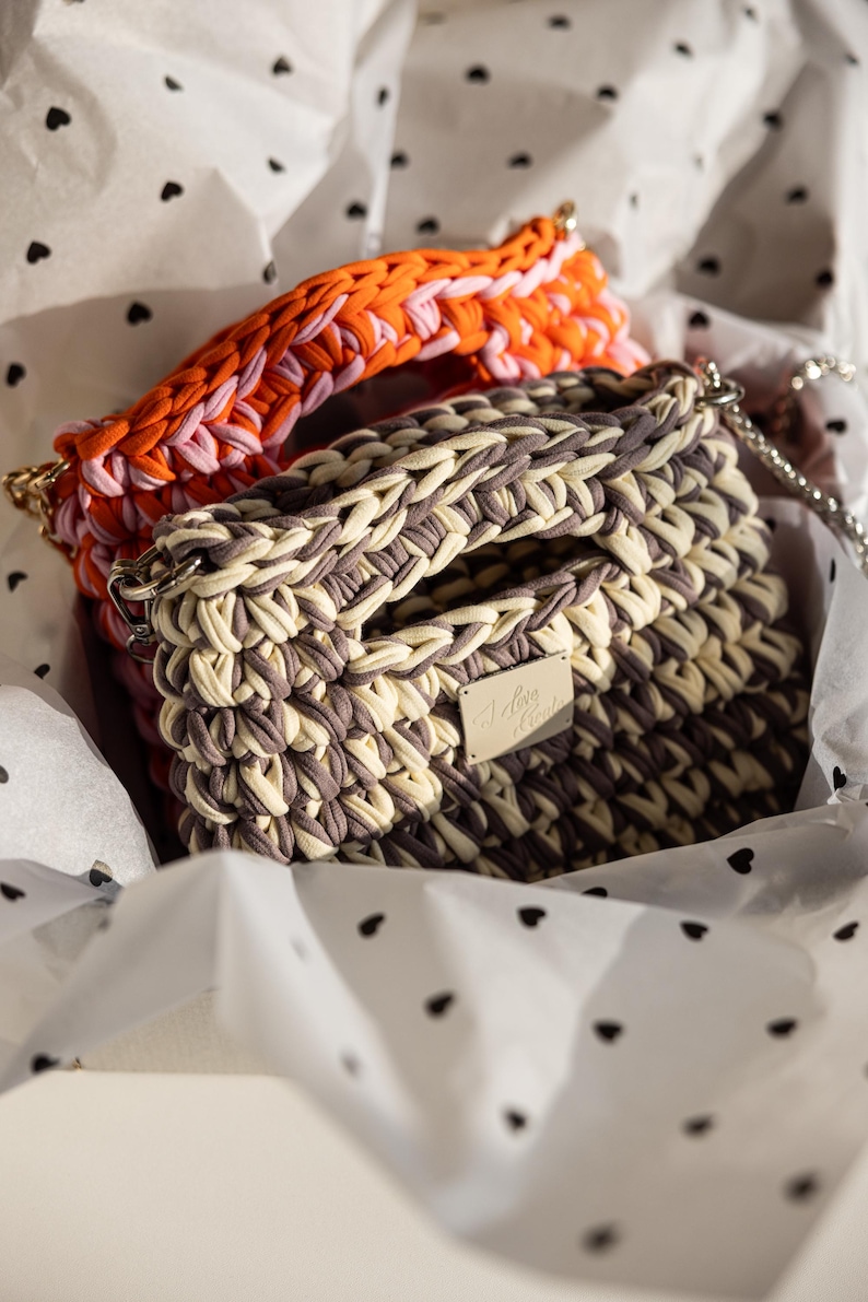 May include: Two handmade crochet bags in a gift box. One bag is a mix of beige and grey, with a silver chain strap and a small metal tag. The other bag has an orange, pink, and white handle. The gift box has a white background with black heart shapes.
