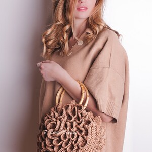 May include: A round, woven handbag with a unique, layered design in a light brown color. The bag features bamboo handles and is held against a neutral-toned outfit. The overall aesthetic is natural and bohemian.