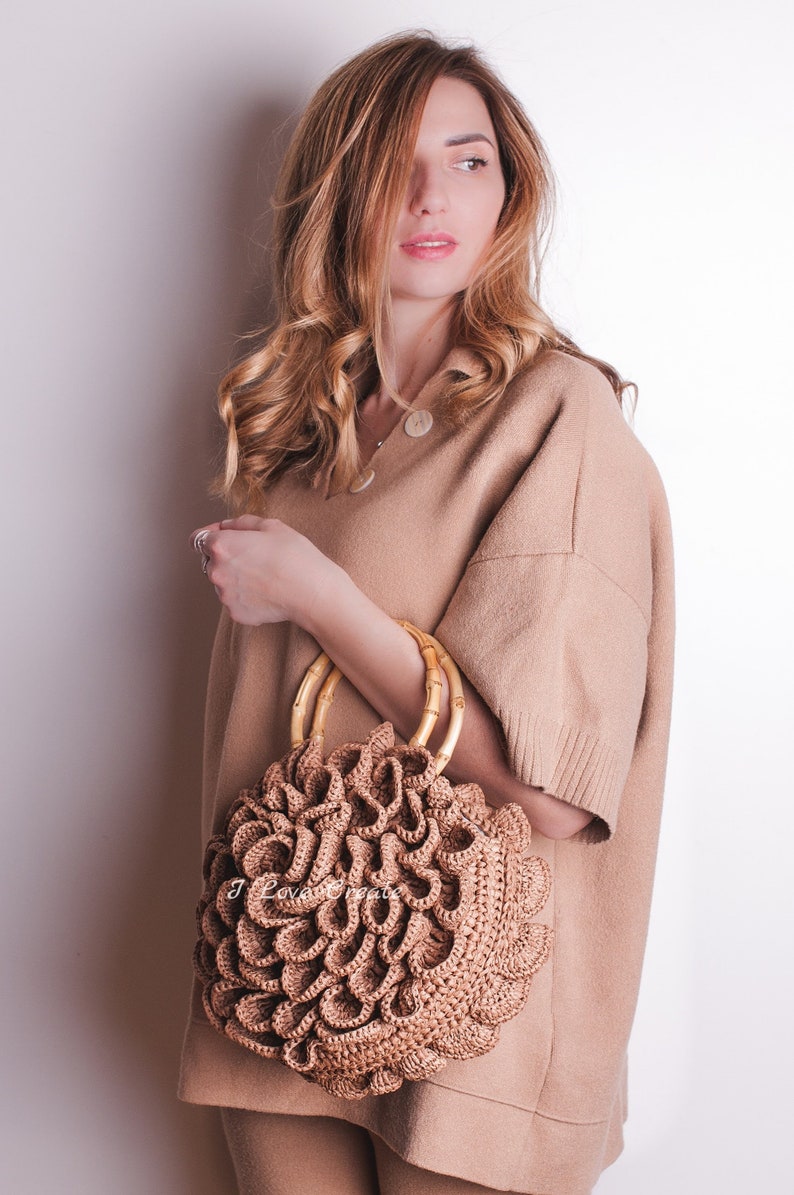 May include: A round, woven handbag with a unique, layered design in a light brown color. The bag features bamboo handles and is held against a neutral-toned outfit. The overall aesthetic is natural and bohemian.