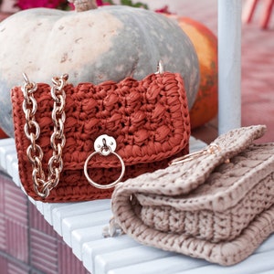 Peut inclure: Un sac à main en crochet de couleur rouille avec une chaîne dorée et un fermoir doré. Un deuxième sac en crochet beige plus petit est à côté. Les sacs sont sur une surface blanche, avec des citrouilles en arrière-plan.