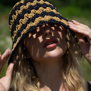 May include: A woman wearing a black and tan crocheted bucket hat. The hat is pulled down low, casting a shadow over her face. She is wearing a silver necklace with a small pendant.