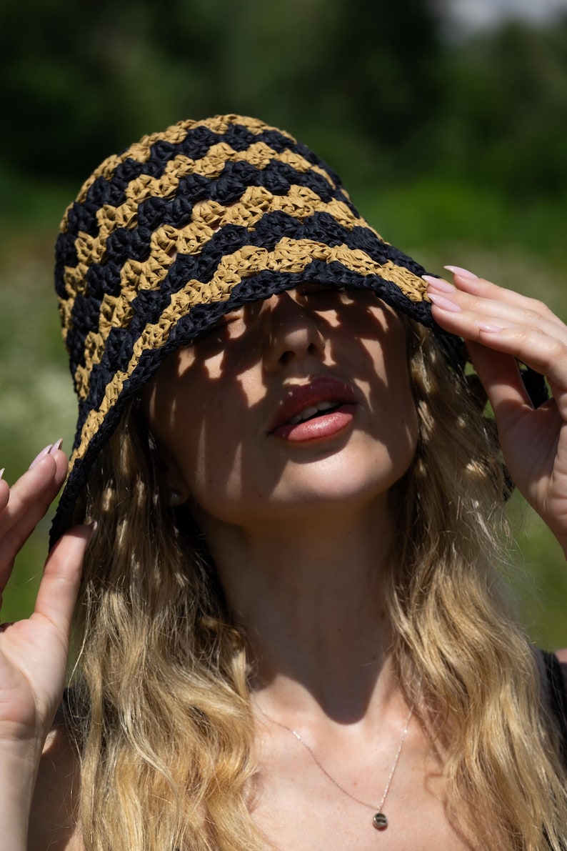 May include: A woman wearing a black and tan crocheted bucket hat. The hat is pulled down low, casting a shadow over her face. She is wearing a silver necklace with a small pendant.