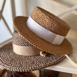Puede incluir: Dos sombreros de paja tejidos a crochet, uno marrón con una cinta blanca y otro beige con una cinta marrón. Los sombreros están apilados uno encima del otro.