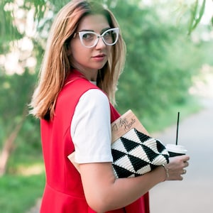 Op de afbeelding: Een persoon draagt een rood vest over een wit shirt en een bril met wit montuur. Ze houdt een zwart-witte geometrische clutch, een houten notitieboek met de tekst "I love Crete" en een koffiebeker met een rietje vast. De achtergrond is een vage groene buitensc&egrave;ne.