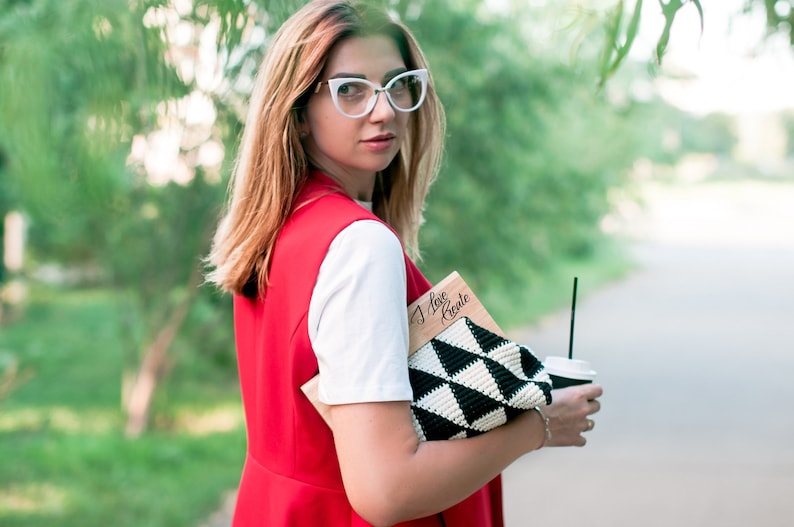 Op de afbeelding: Een persoon draagt een rood vest over een wit shirt en een bril met wit montuur. Ze houdt een zwart-witte geometrische clutch, een houten notitieboek met de tekst "I love Crete" en een koffiebeker met een rietje vast. De achtergrond is een vage groene buitensc&egrave;ne.
