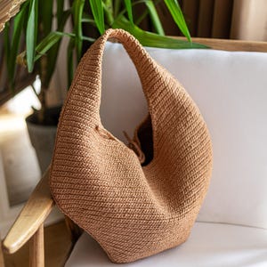 May include: A tan crochet shoulder bag with a rounded shape and a single strap. The bag is made of a textured, woven material and sits on a white chair. The bag's interior is visible, and the background includes a green plant.
