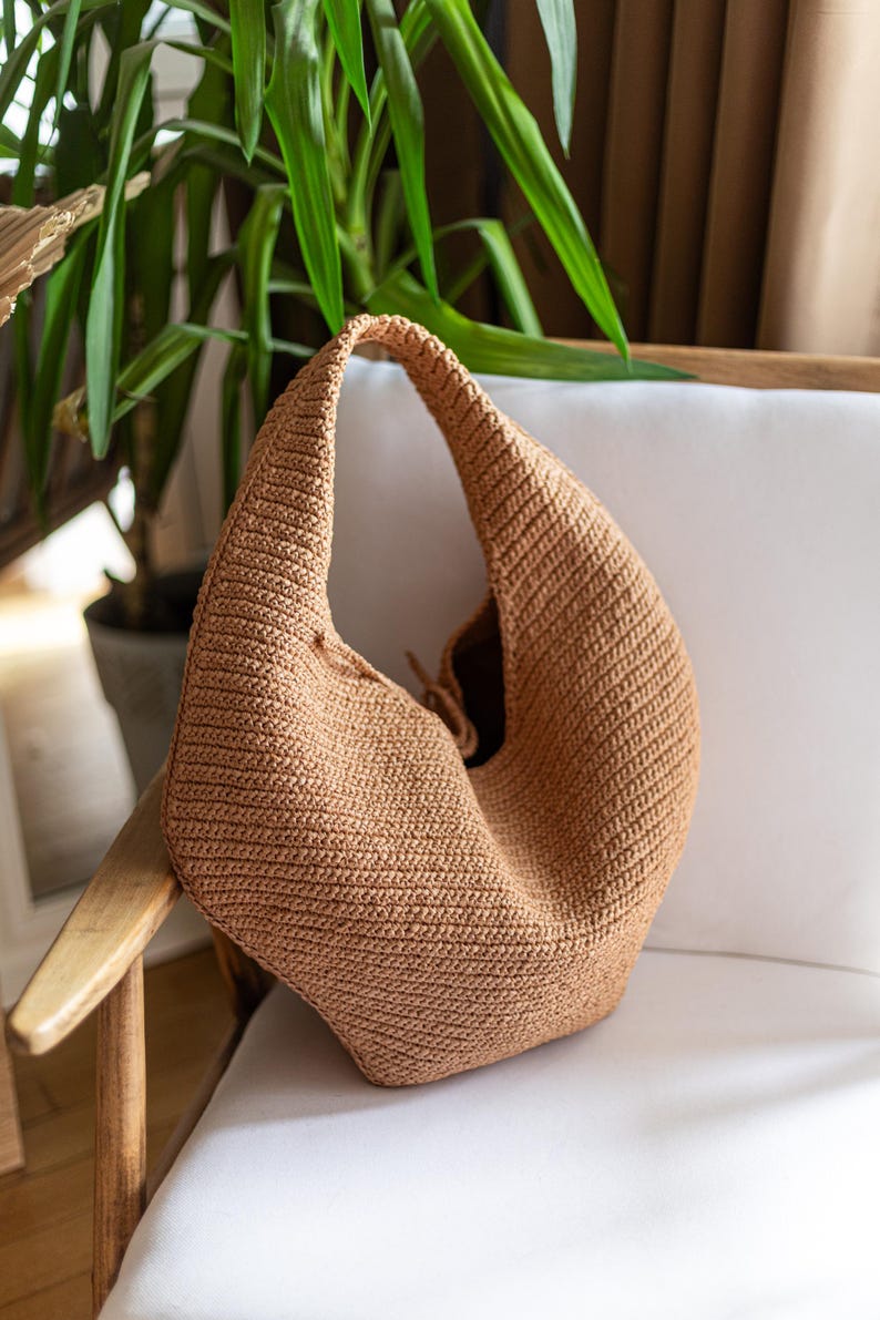 May include: A tan crochet shoulder bag with a rounded shape and a single strap. The bag is made of a textured, woven material and sits on a white chair. The bag's interior is visible, and the background includes a green plant.