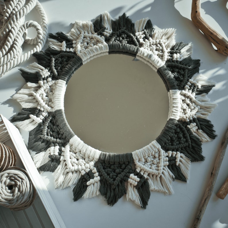 May include: A round mirror with a macrame frame in black and white. The frame is made of thick rope and has a fringe around the edge.
