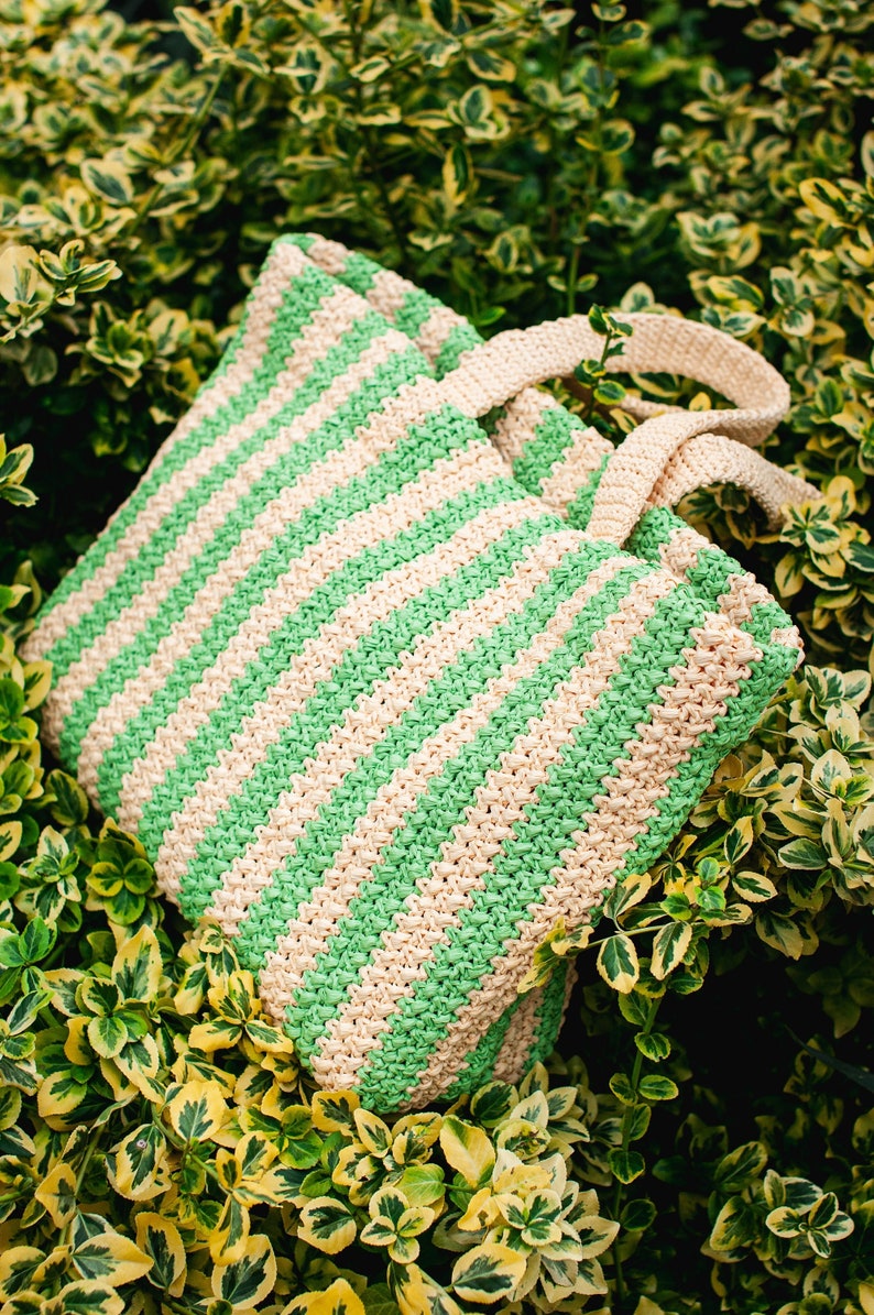 May include: A green and beige striped tote bag with matching handles rests among green and yellow foliage. The bag is made of a woven material, and the stripes run diagonally across the bag.