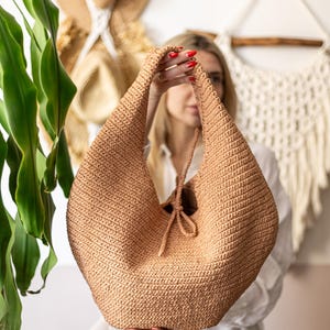 May include: A tan crochet hobo bag with a drawstring closure. The bag is held up by the handles. The bag is made of a woven material. The background includes hats and a macrame wall hanging.