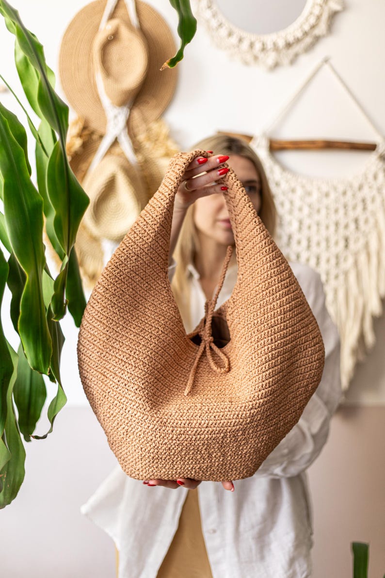 May include: A tan crochet hobo bag with a drawstring closure. The bag is held up by the handles. The bag is made of a woven material. The background includes hats and a macrame wall hanging.