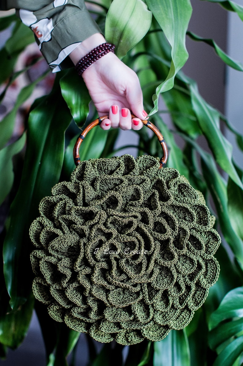 May include: A handcrafted, olive green crochet bag with a unique, textured design. The bag features a bamboo handle with dark accents. The bag is held in front of a backdrop of green foliage.