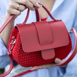 Könnte beinhalten: Eine rote Handtasche mit einem strukturierten, gehäkelten Körper und einer glatten, einfarbigen roten Klappe. Die Tasche verfügt über einen Tragegriff und einen abnehmbaren Schulterriemen aus passendem rotem Material, befestigt mit silberfarbenen Metallbeschlägen.
