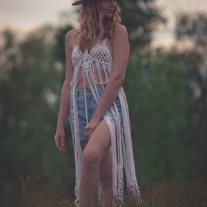 Peut inclure: Une femme portant un haut en crochet blanc avec des d&eacute;tails en frange, un short en jean bleu et un chapeau de paille brun. Elle se tient dans un champ d'herbe haute.