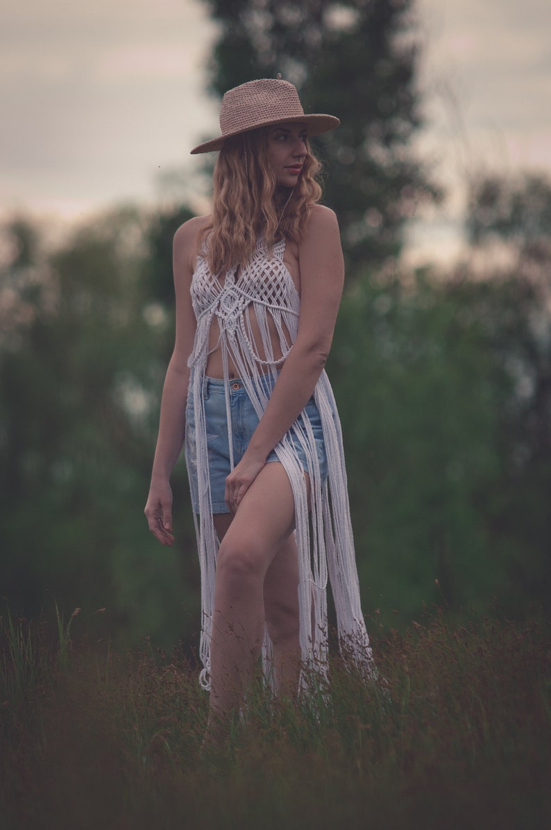 Peut inclure: Une femme portant un haut en crochet blanc avec des d&eacute;tails en frange, un short en jean bleu et un chapeau de paille brun. Elle se tient dans un champ d'herbe haute.
