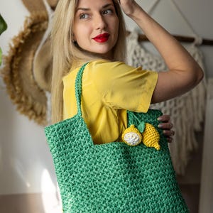 May include: A green crocheted tote bag with a lemon applique. The bag has a rectangular shape with a handle. The lemon applique is yellow and white. The bag is held by a person wearing a yellow shirt.
