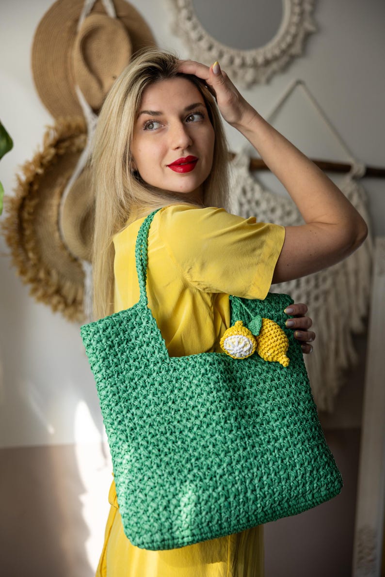 May include: A green crocheted tote bag with a lemon applique. The bag has a rectangular shape with a handle. The lemon applique is yellow and white. The bag is held by a person wearing a yellow shirt.