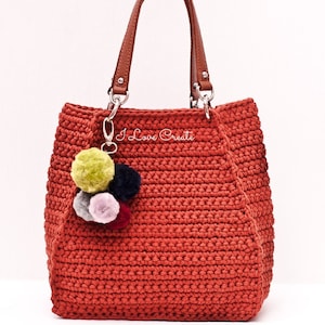 May include: A red crocheted tote bag with brown leather handles and a silver chain with pom-pom keychains. The bag has the text "I Love Create" embroidered on the front.