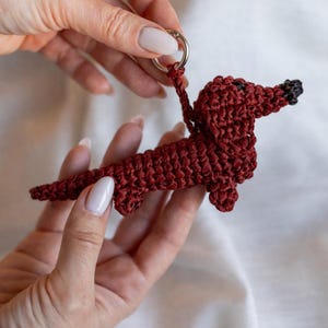 Könnte beinhalten: Ein handgefertigter, roter Dackel-Schlüsselanhänger aus Häkelarbeit. Der Hund hat einen langen Körper, kurze Beine und eine schwarze Nase. Der Schlüsselanhänger ist ein charmantes Accessoire.