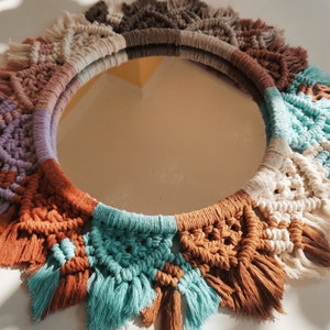 May include: A round mirror with a macrame frame in shades of brown, white, and blue. The frame is made of knotted cords and has a fringe around the edge.