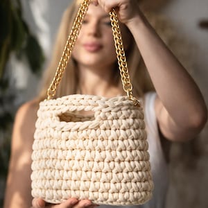 May include: Cream-colored crochet handbag with a gold chain strap. The bag has a rectangular shape with a handle opening at the top. The chain is thick and gold-toned, contrasting with the neutral color of the bag.