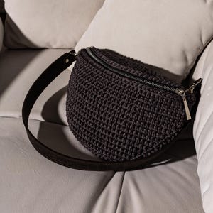 May include: A dark gray crocheted fanny pack with a black adjustable strap. The bag has a zipper closure and a silver zipper pull.