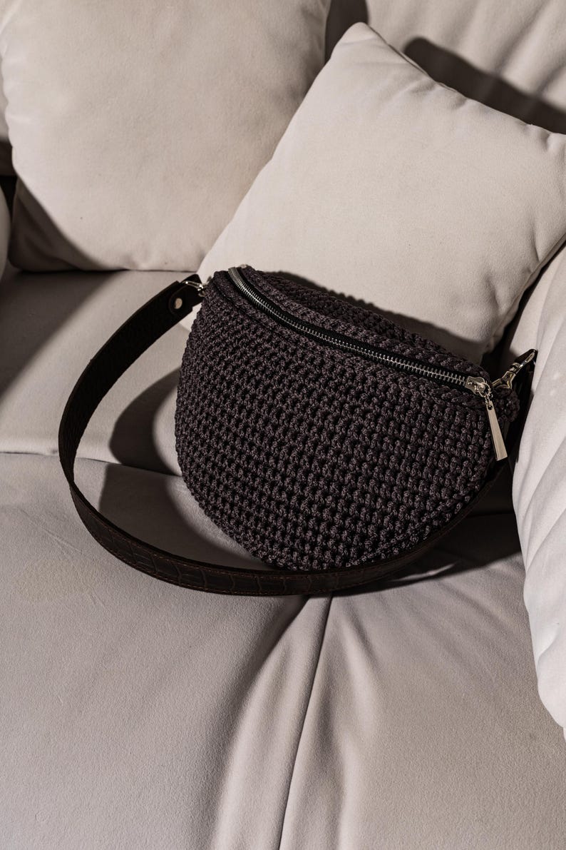 May include: A dark gray crocheted fanny pack with a black adjustable strap. The bag has a zipper closure and a silver zipper pull.
