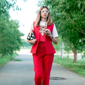 Op de afbeelding: Een vrouw in een rode broekpak, wit grafisch T-shirt en rode schoenen met transparante hakken loopt over een pad. Ze draagt een zwart-witte clutch en een koffiekopje. Het vest en de broek zijn felrood.