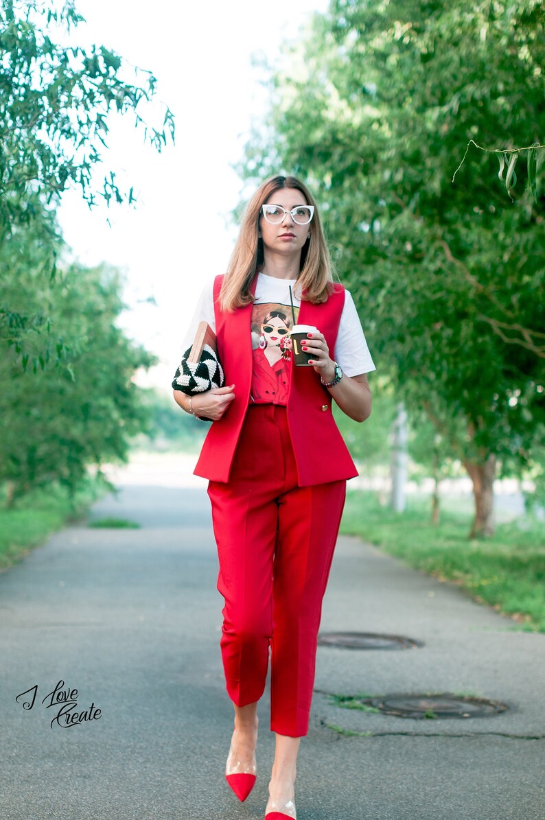 Op de afbeelding: Een vrouw in een rode broekpak, wit grafisch T-shirt en rode schoenen met transparante hakken loopt over een pad. Ze draagt een zwart-witte clutch en een koffiekopje. Het vest en de broek zijn felrood.