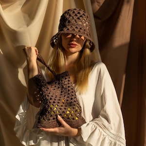 May include: A woman wearing a brown crocheted sun hat and a white crochet bag with a brown strap. The bag is filled with green produce. She is standing in front of a beige and brown fabric backdrop.