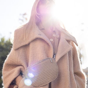 May include: A woman wearing a light brown coat with a large collar and a crocheted beige fanny pack worn as a crossbody bag.