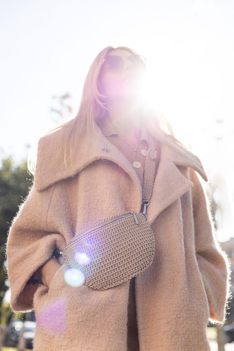 May include: A woman wearing a light brown coat with a large collar and a crocheted beige fanny pack worn as a crossbody bag.