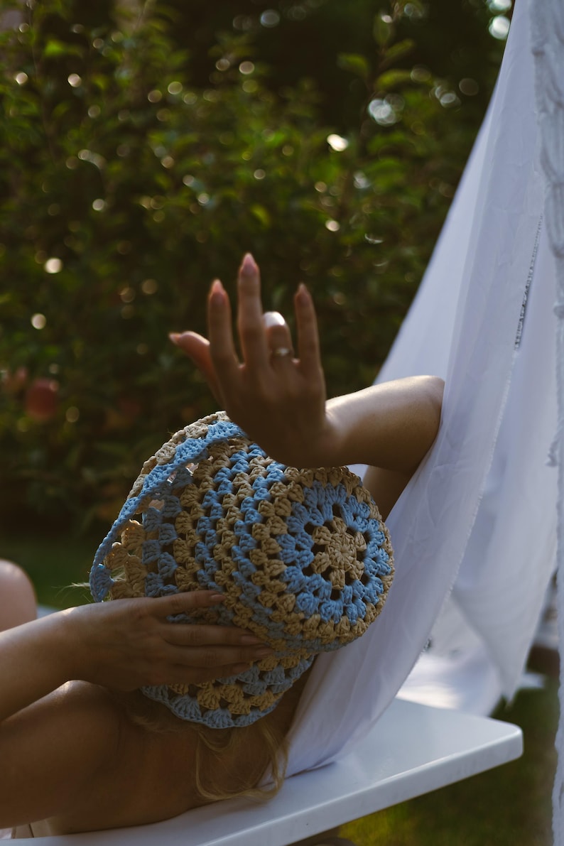 May include: A woman wearing a blue and brown crocheted sun hat while relaxing in a hammock.