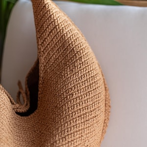 May include: A tan-colored, crocheted shoulder bag with a rounded shape. The bag has a single strap and a textured surface. The bag is made of a woven material and appears to be handmade.