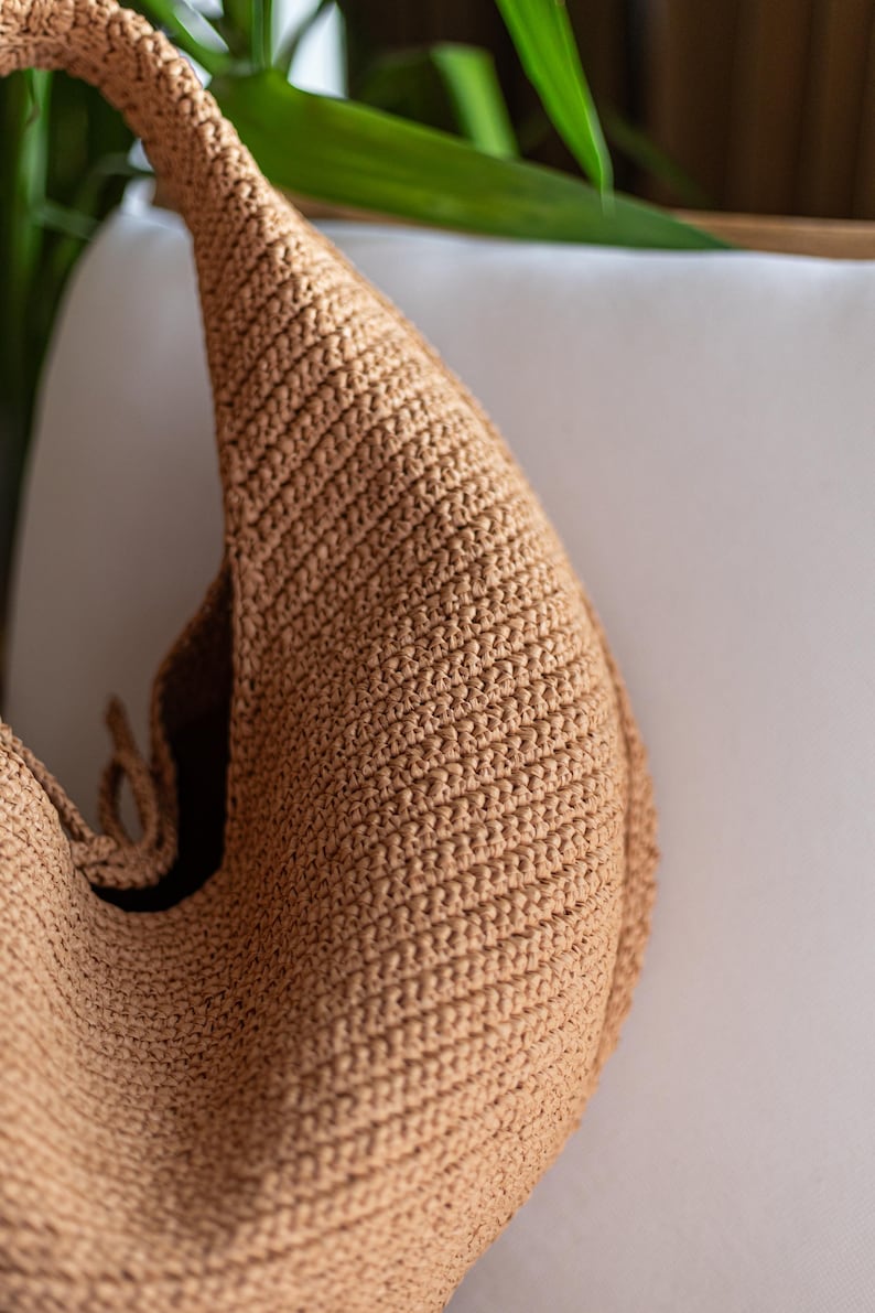May include: A tan-colored, crocheted shoulder bag with a rounded shape. The bag has a single strap and a textured surface. The bag is made of a woven material and appears to be handmade.
