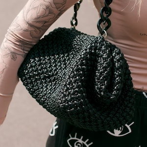 Può includere: Una borsa a mano nera intrecciata con una spessa tracolla a catena di plastica nera. La borsa è realizzata in un materiale intrecciato fitto e ha una forma arrotondata leggermente gonfia.