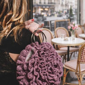 May include: A mauve-colored, crocheted handbag with a unique, textured design. The bag features two silver metal handles. The background shows a cafe setting with tables and chairs, suggesting a stylish accessory.