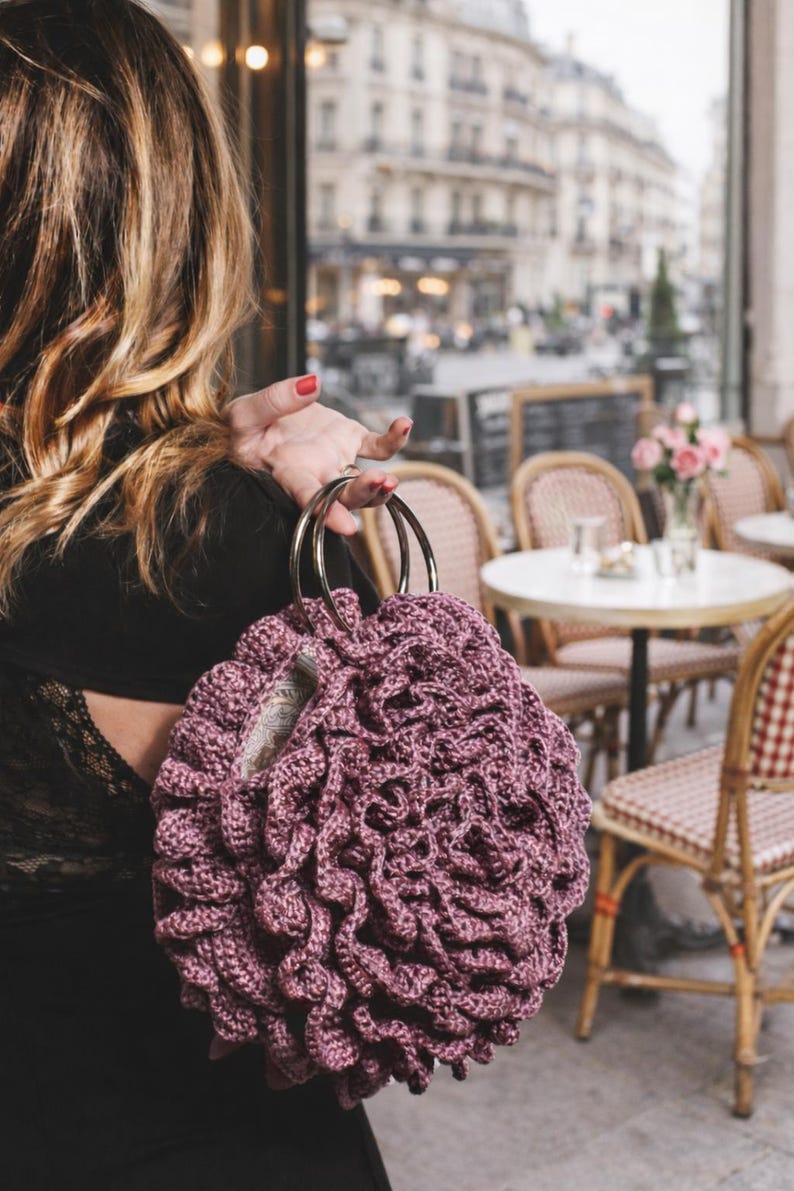 May include: A mauve-colored, crocheted handbag with a unique, textured design. The bag features two silver metal handles. The background shows a cafe setting with tables and chairs, suggesting a stylish accessory.