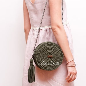 Puede incluir: Un bolso redondo de crochet verde con una etiqueta de cuero marrón que dice "I Love Create". El bolso tiene una larga correa de cadena plateada y un borla verde.