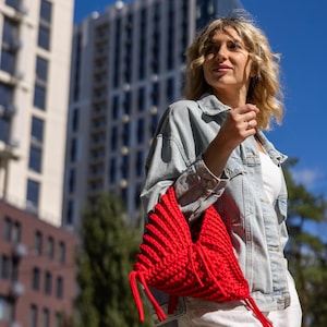 May include: A bright red, handmade crochet bag with long, matching drawstrings. The bag is held by a person wearing a light blue denim jacket and white pants. The background features tall buildings and a clear blue sky.