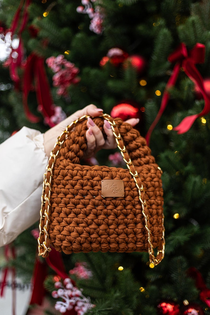Op de afbeelding: Een bruine gehaakte handtas met een gouden kettingband. De tas heeft een klein leren label met de tekst "I love croche" erop.