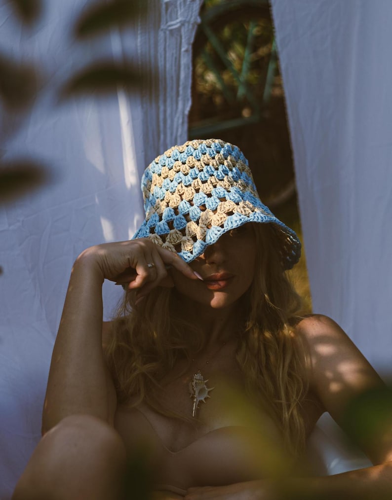 May include: A woman wearing a blue and beige crochet bucket hat, with her hair covering her eyes. She is wearing a seashell necklace and a light brown top.