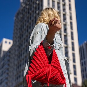 May include: A vibrant red, handmade knitted bag with a unique, textured design. The bag features long, dangling red cords and is carried over the shoulder. The background includes a modern building and a clear blue sky.