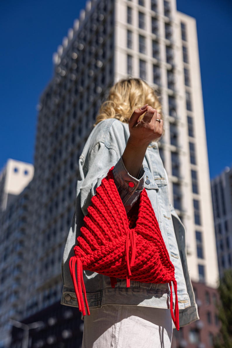 May include: A vibrant red, handmade knitted bag with a unique, textured design. The bag features long, dangling red cords and is carried over the shoulder. The background includes a modern building and a clear blue sky.