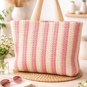May include: A large, rectangular crochet tote bag with vertical stripes in pink and cream. The bag has two cream-colored handles and sits on a woven mat. Pink sunglasses and a notebook are in the foreground.