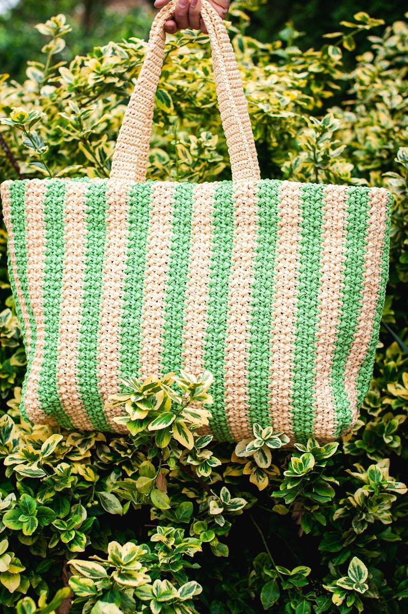 May include: A crocheted tote bag with alternating vertical stripes of light green and beige. The bag has matching beige handles and is set against a backdrop of green and yellow foliage.